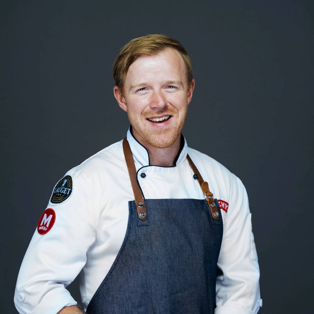 En person iført hvit kokkejakke og blå forkle, med navn Petter Johannessen, mot en mørk bakgrunn.