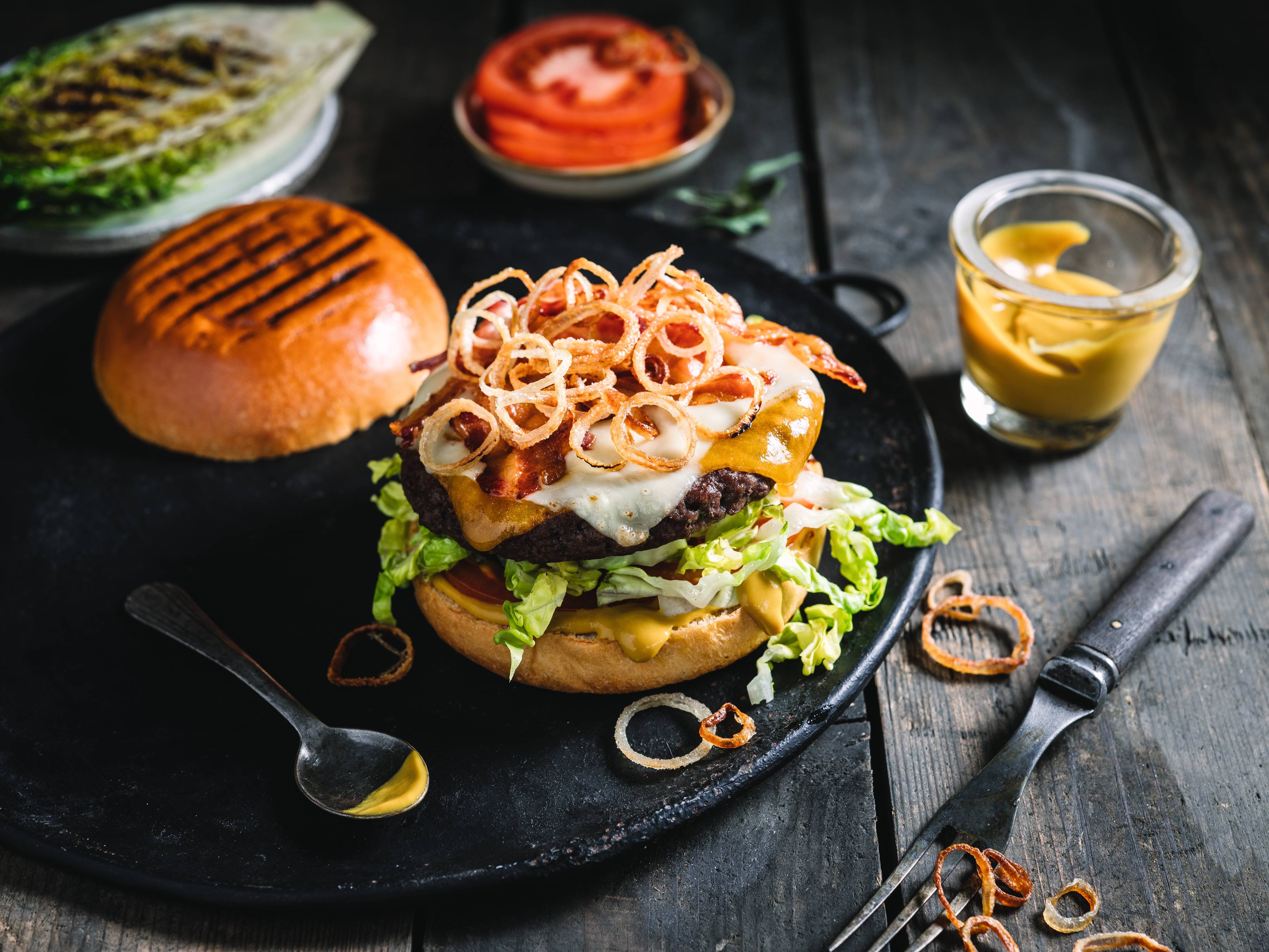 En klassisk burger med kjøtt, salat, ost og løk på en svart stekeplate, med en liten skål med ketchup og en glass med sitronvann ved siden av.