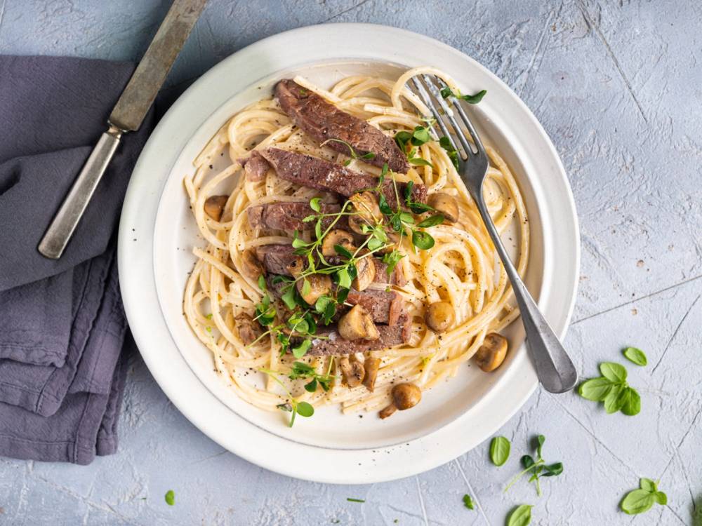 Entrecote med pasta og grønne urter på en hvit tallerken, med bestikk ved siden av.