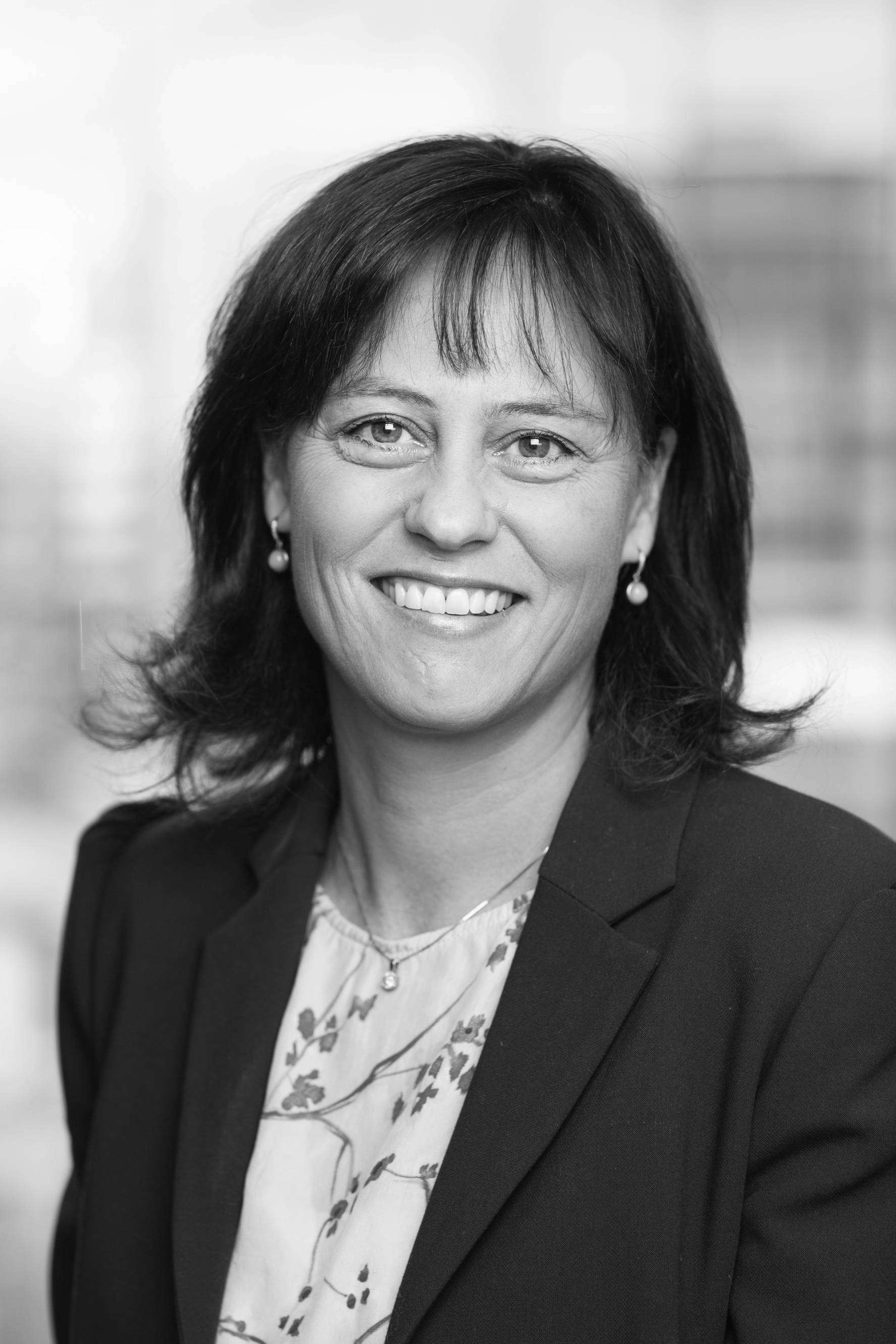 Portrett av en kvinne med mørkt, skulderlangt hår iført blazer og blomstret bluse, uten ansikt synlig på grunn av ansiktsdekning.