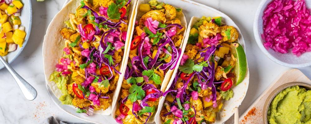 To tacos fylt med blomkål, kål, og grønnsaker, ser ut til å være på et fat med tilbehør i små boller rundt.