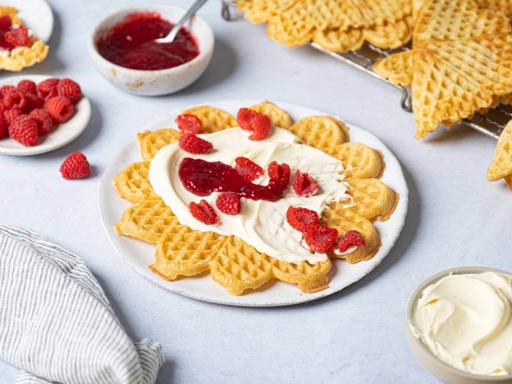 Vafler med rømme og bringebær på en hvit tallerken, omgitt av flere vaffelplater og en skål med bringebær.