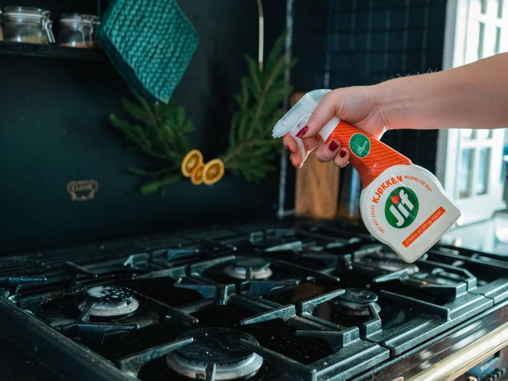 En person sprayer rengjøringsmiddel på en komfyr i et kjøkken, med grønne planter i bakgrunnen.