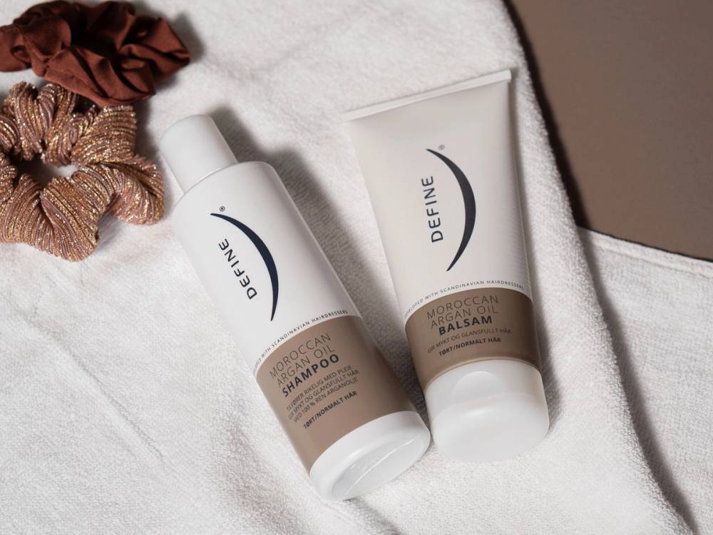 To hårpleieprodukter, shampoo og balsam, ligger på en hvit håndkle med en brun hårstrikk ved siden av.