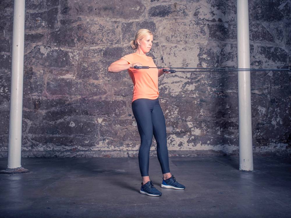 En person står oppreist og holder en treningsstang foran brystet i et treningsrom.