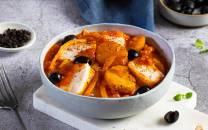 Bacalao med poteter, svarte oliven og en tomatsaus i en hvit oval serveringsskål.