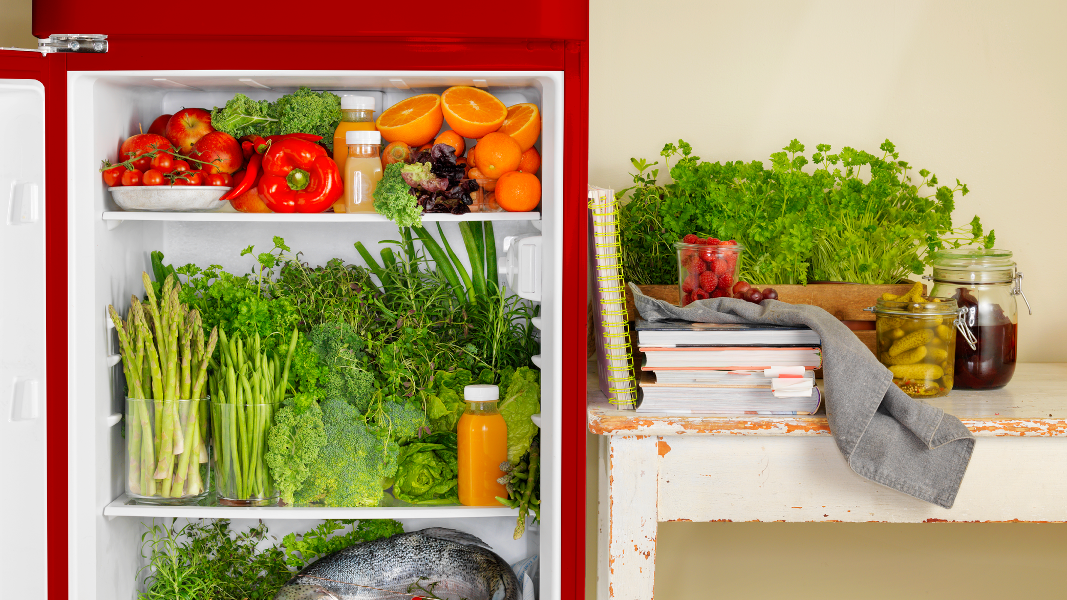 Et kjøleskap fylt med frukt, grønnsaker og juice, plassert ved siden av en benk med urter og matvarer.