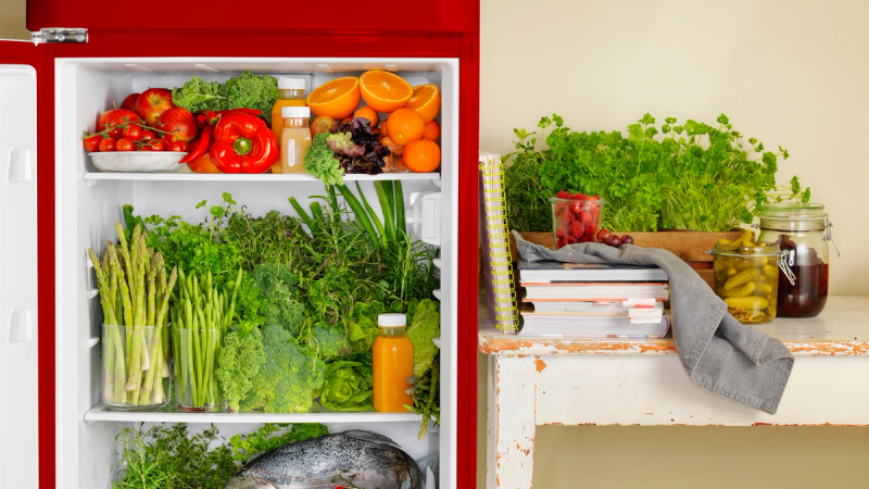 Et kjøleskap fylt med frukt, grønnsaker og juice, plassert ved siden av en benk med urter og matvarer.