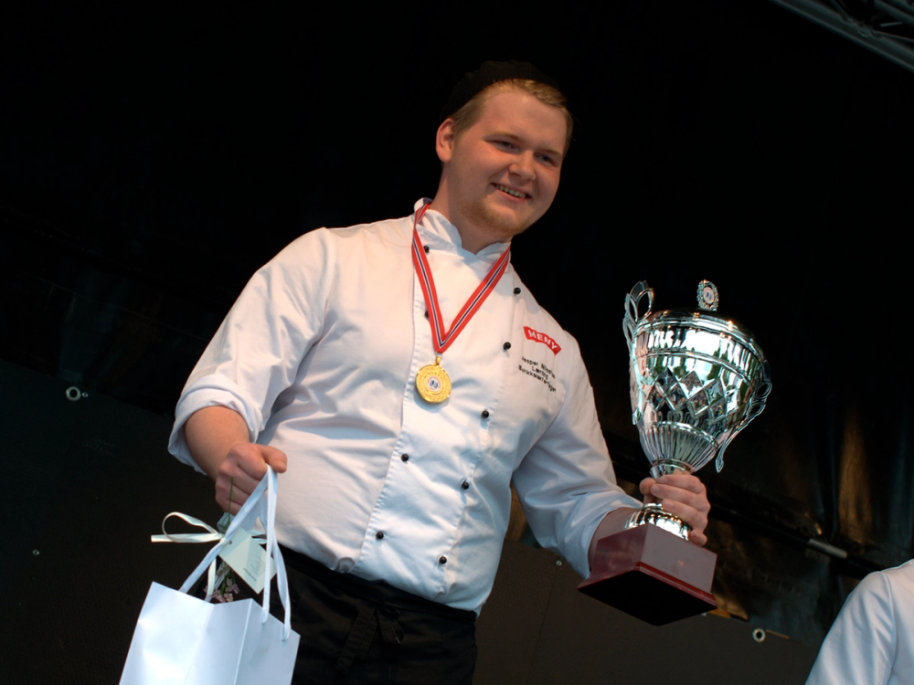 En person i hvit kokkeflate holder en stor pokal og en gavepose, sannsynligvis etter en konkurranse eller prisutdeling.