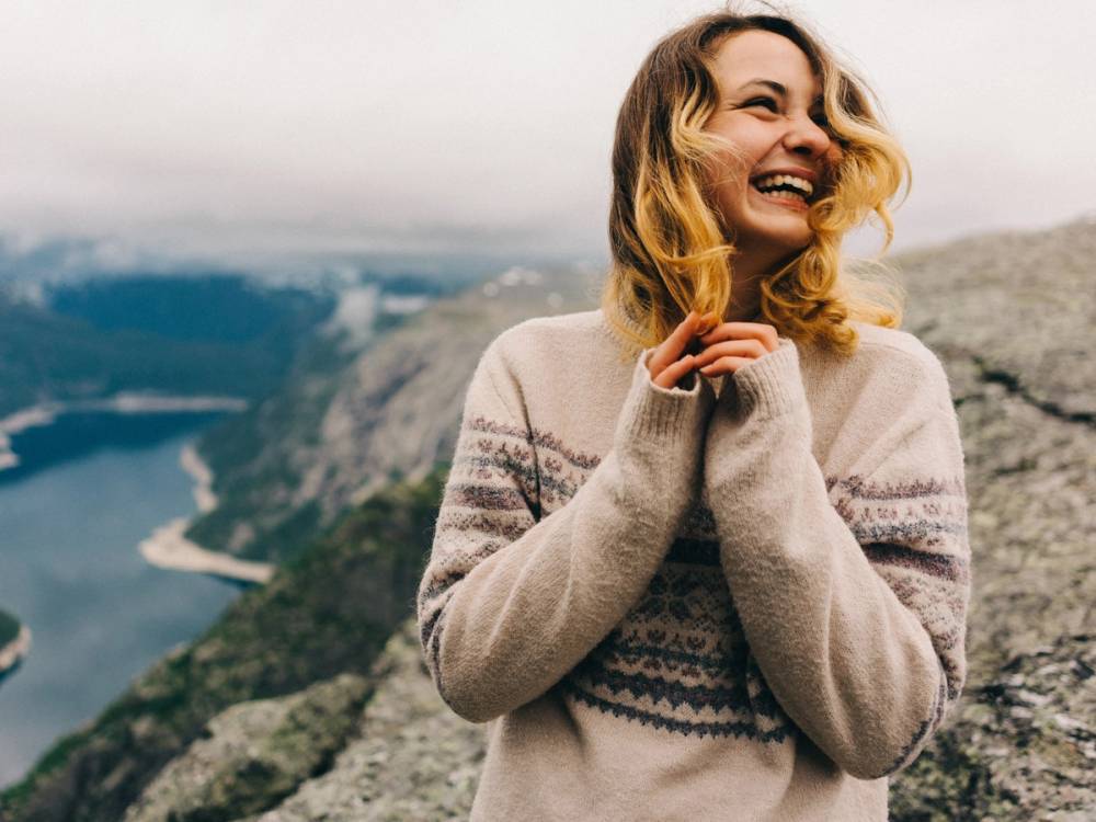 En person med blonder hår står på en fjelltopp med utsikt over en dal og en elv under en overskyet himmel.