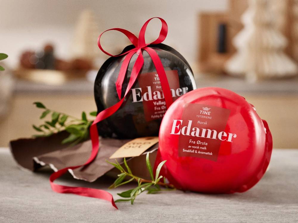 To runde Edamer-oster, en svart med rød sløyfe og en rød uten sløyfe, plassert på et bord med grønne blader rundt.