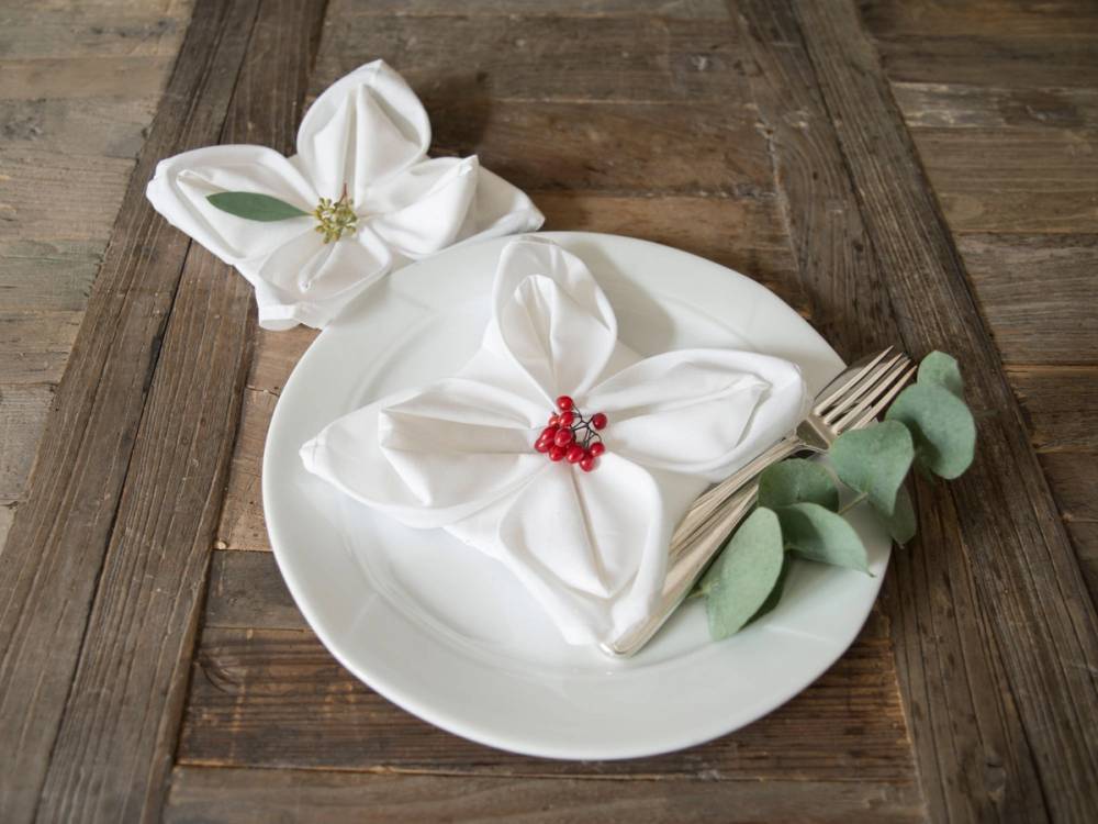 Servietter med julemotiv brettet som blomster på et hvitt fat, pyntet med røde bær og grønne blader, på et trebord.