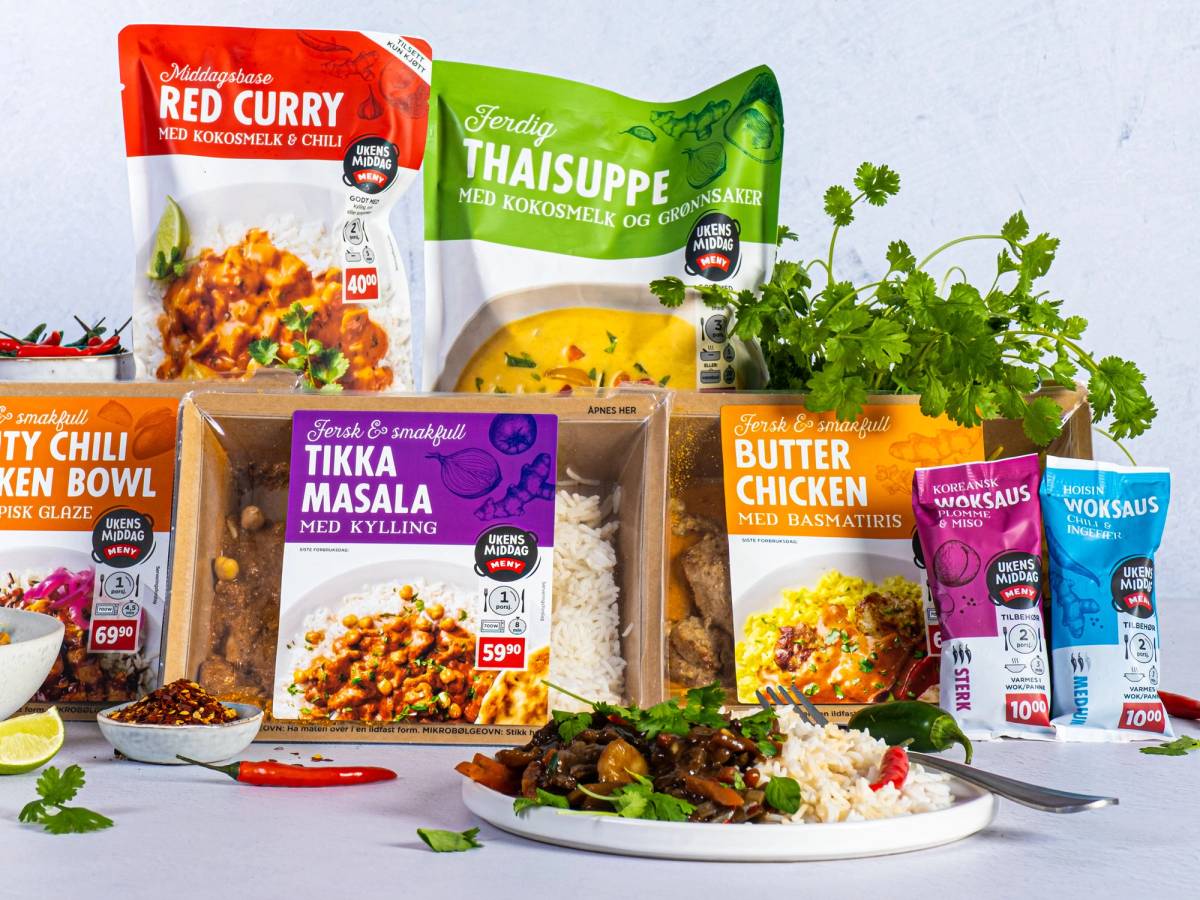 En rekke krydderposer, en pakke med thaisuppe, en pakke med tikkamasala, en pakke med butter chicken, og fersk koriander på et kjøkkenbord med en tallerken med ferdiglaget middag.