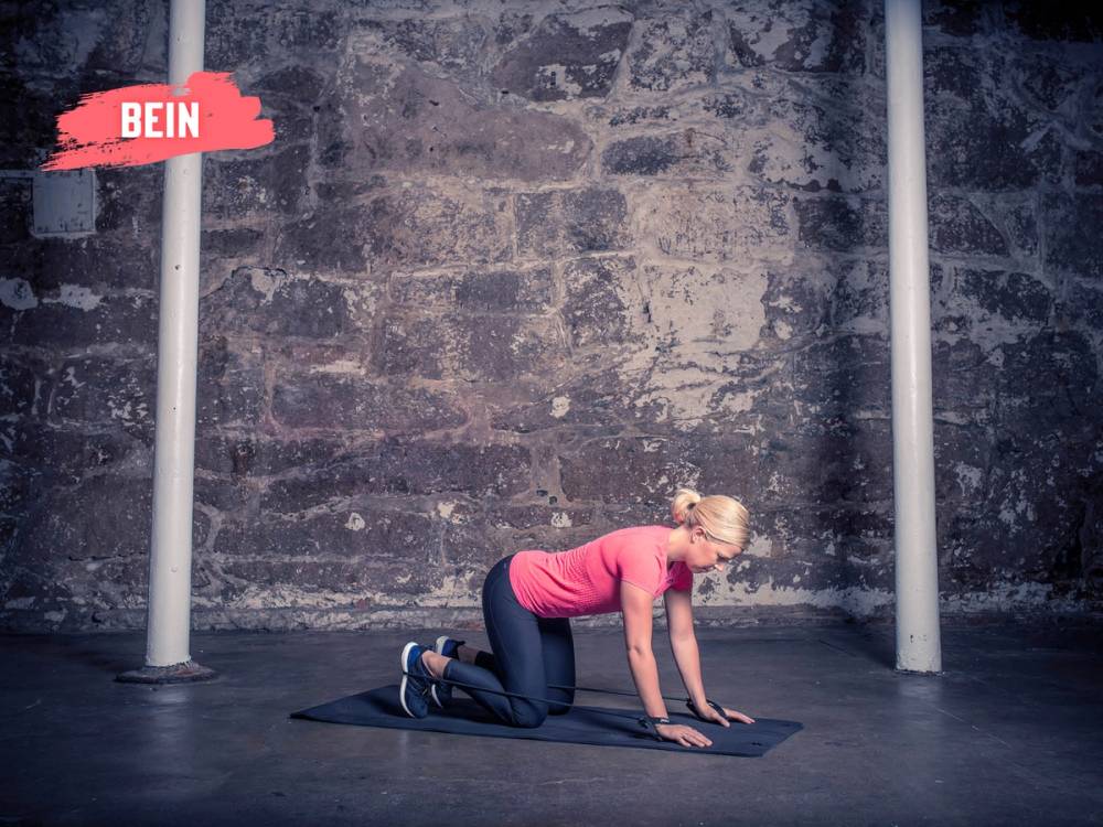 En kvinne i rosa treningstøy som gjør en øvelse på gulvet i et treningsrom med en murvegg i bakgrunnen.