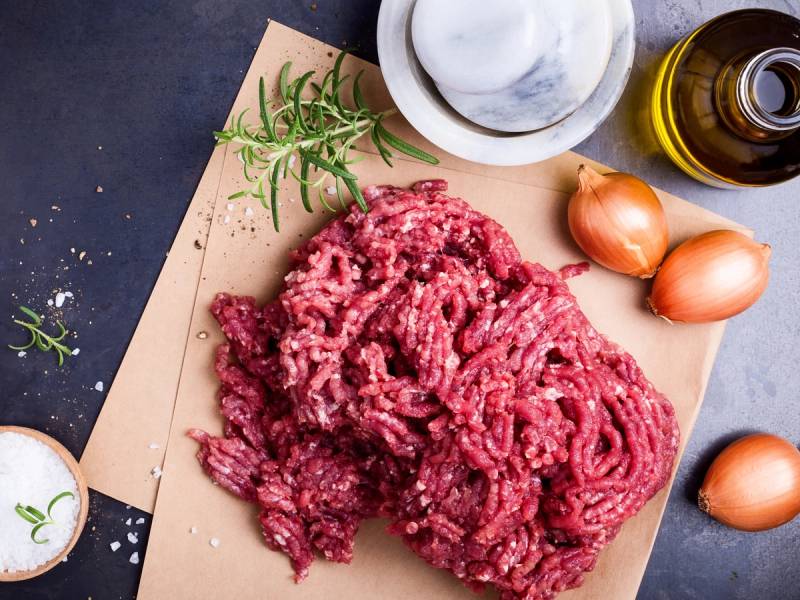 Kjøttdeig på et skjærebrett med løk, urter og krydder ved siden av, klar til å bli brukt i matlaging.