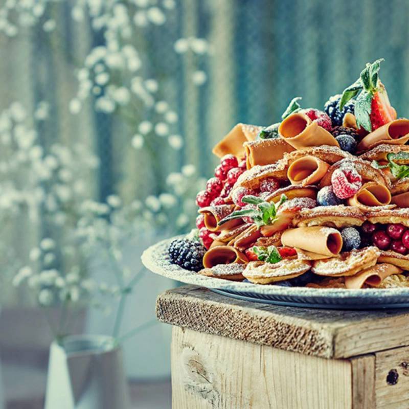 En tallerken med en dekorativ dessert med frukt, bær og sprøytetopping, plassert på et trebord med en blomsterbakgrunn.