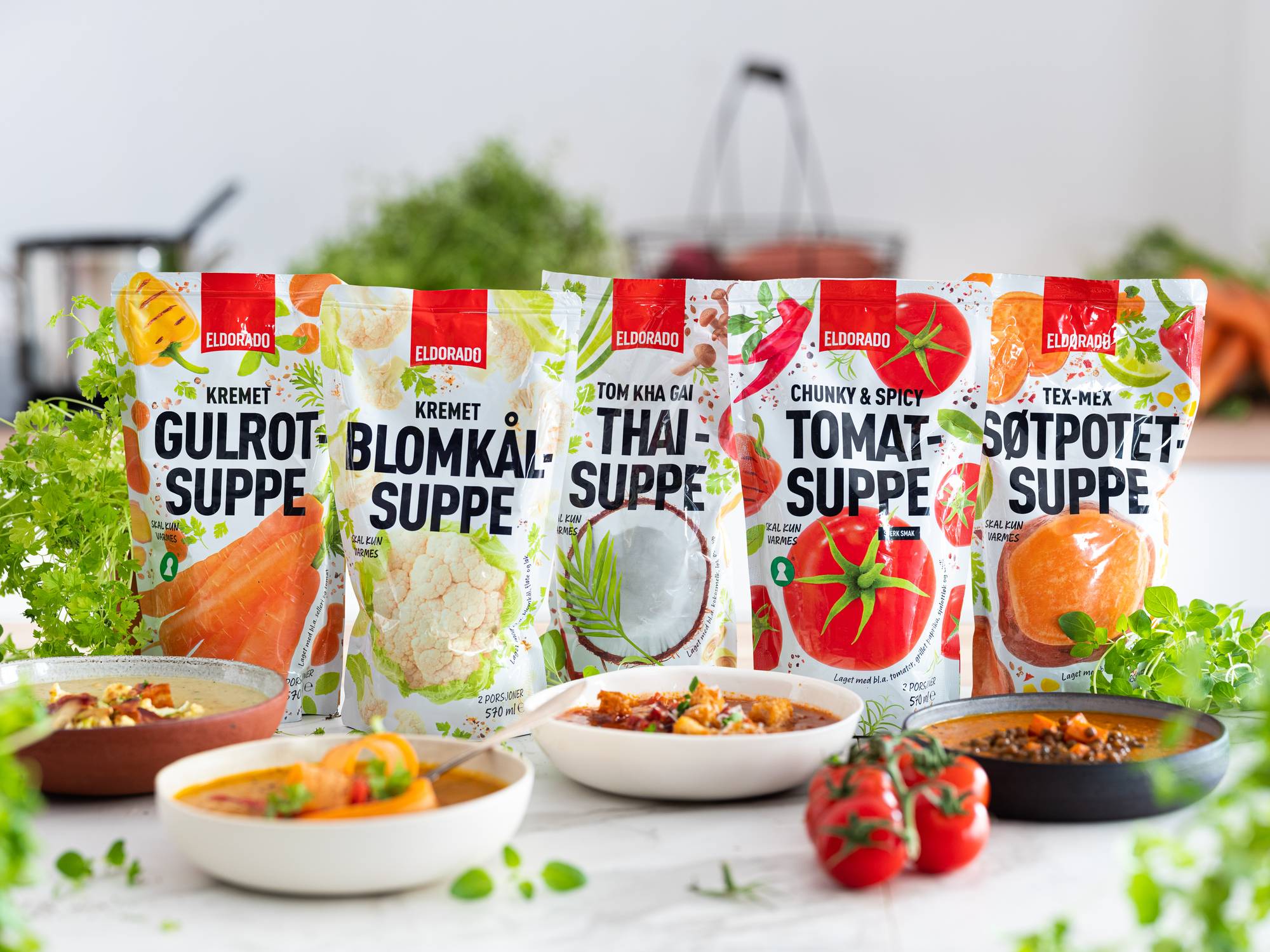 Fremstående pakker med ulike supper, inkludert gulrotsuppe, blomkåpsuppe, thaisuppe, tomatsuppe og søtpotetsuppe, med noen ferdige supper i skåler foran.