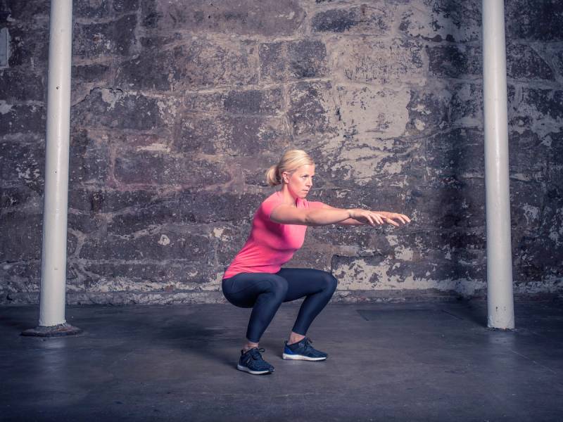En person i rosa treningsklær gjør en knebøy i et treningsrom med en murvegg i bakgrunnen.