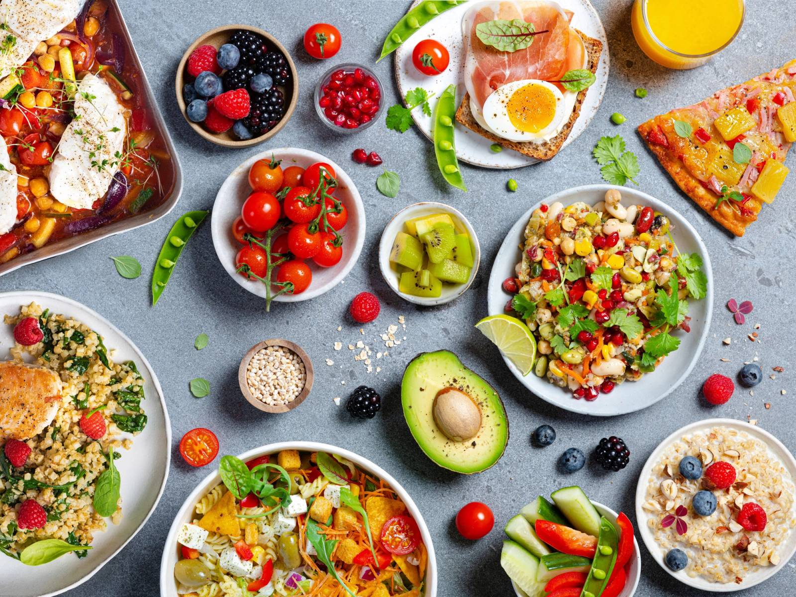 Et utvalg av sunne frokostretter og frukt på et bord, inkludert salater, avokado, egg, og frukt, med en glass med appelsinjuice i bakgrunnen.