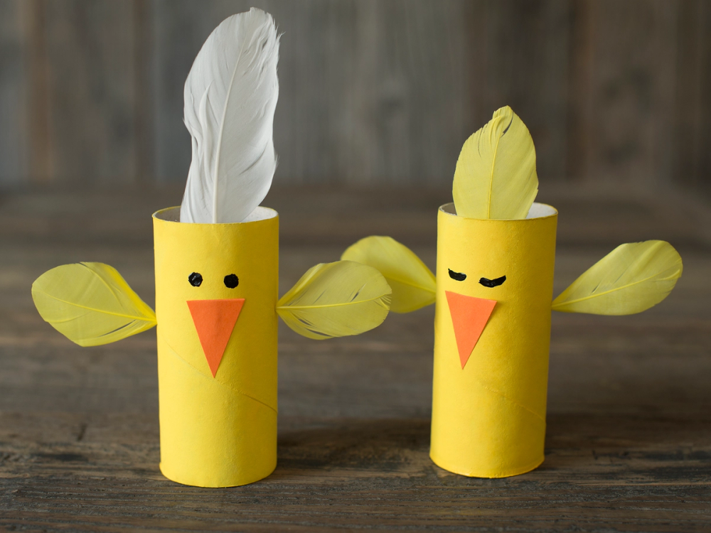 To dorullkyllinger laget av papir med fjær og papirvinger, en med en hvit fjær og den andre med en gul fjær, plassert på et treverk.