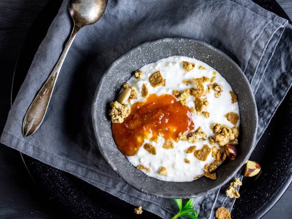 Cottage cheese med granola og honning i en grå bolle på et svart underlag med en skje og en serviett ved siden av.