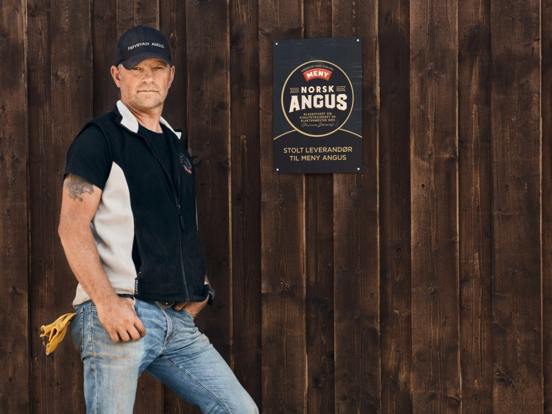 Person står foran en trevegg med en Angus-plate på, iført en svart og hvit skjorte og jeans, med en gressklipper i hånden.