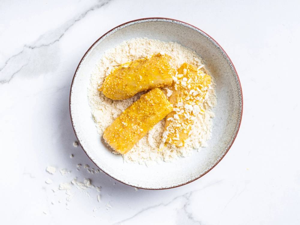 Fiskepinner dekket med mel og ost, klar for steking, sett i en bolle på et hvitt underlag.