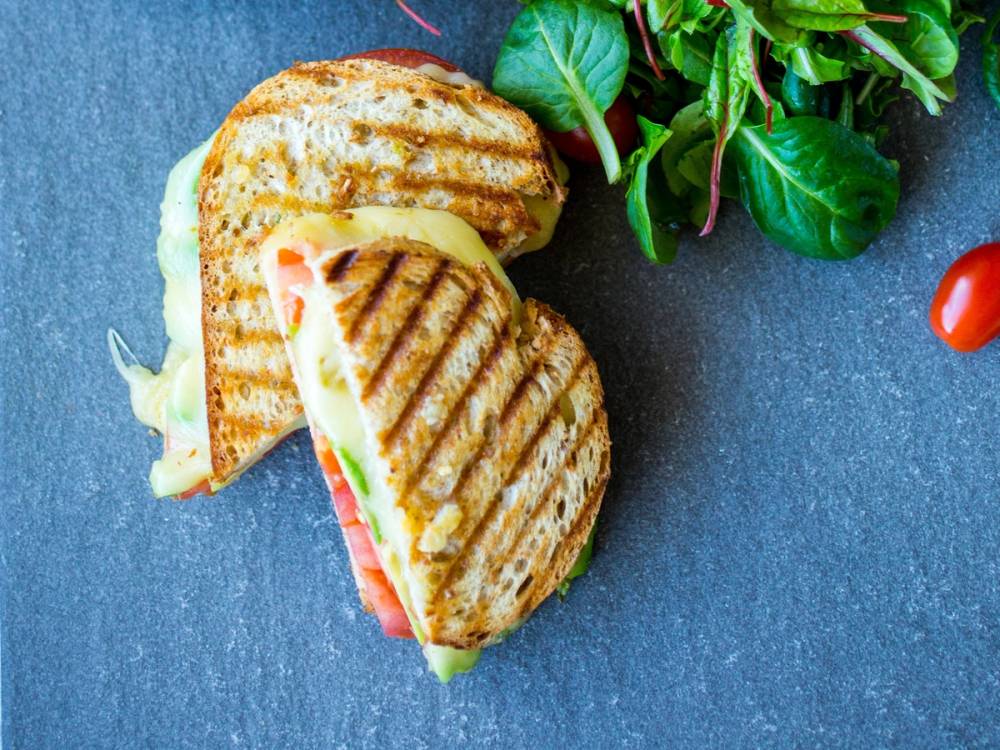 To grillede paninier med ost og skinke på en blå bakgrunn, med en grønn salat og cherrytomater i nærheten.