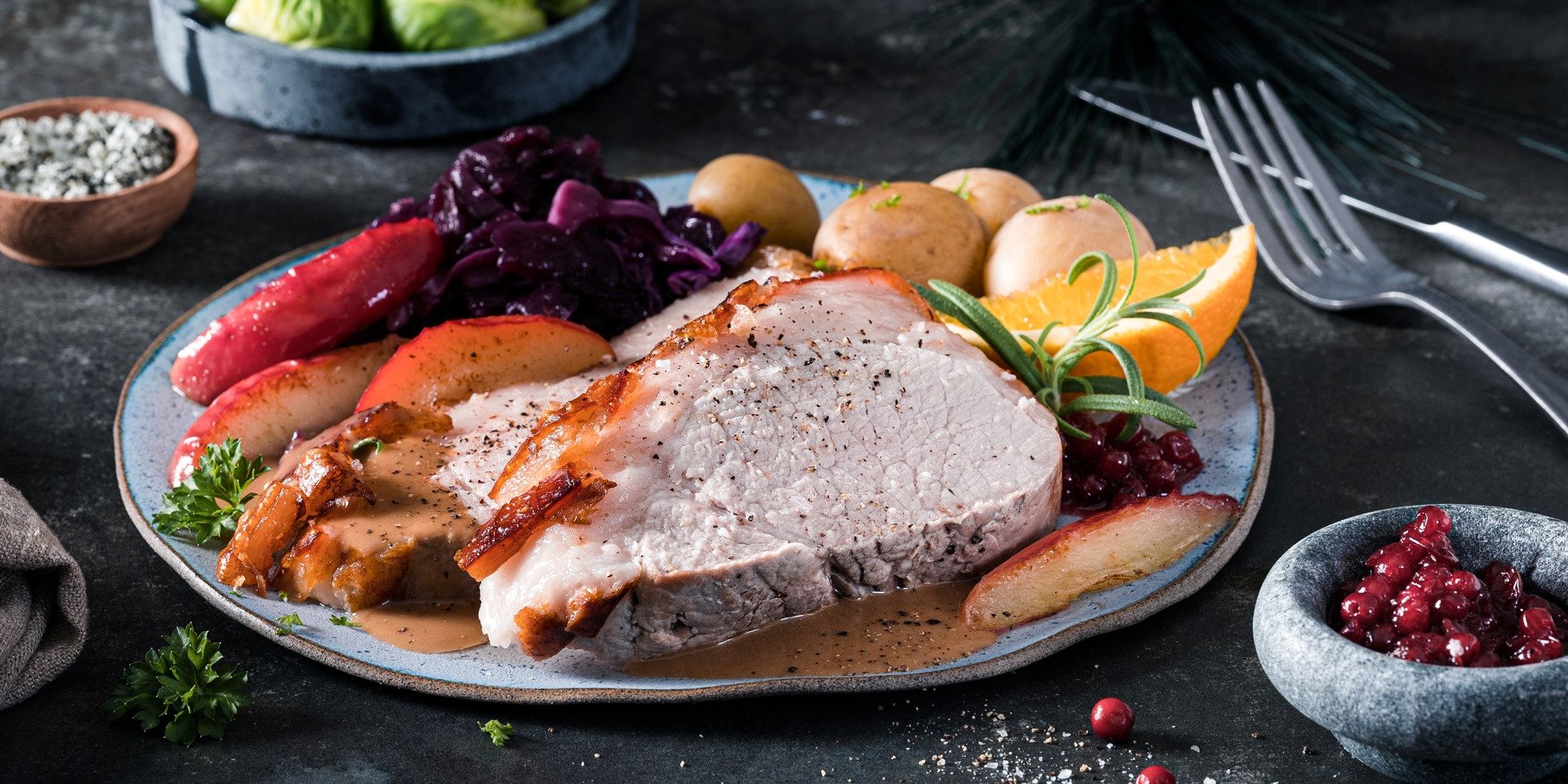 Skinkestek med ben servert på en tallerken sammen med poteter, grønnsaker og saus, omgitt av bær og sitronbåter.