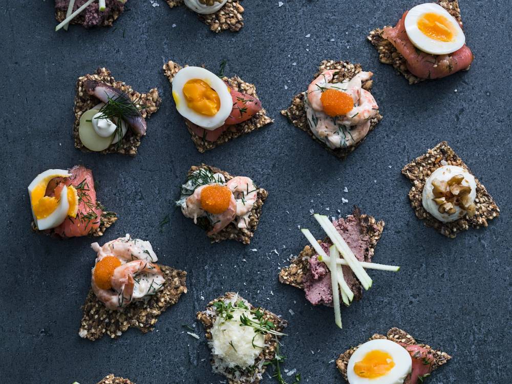 Små åpne kaker med ulike pålegg og pynt, noen med egg og grønnsaker, på en mørk overflate.