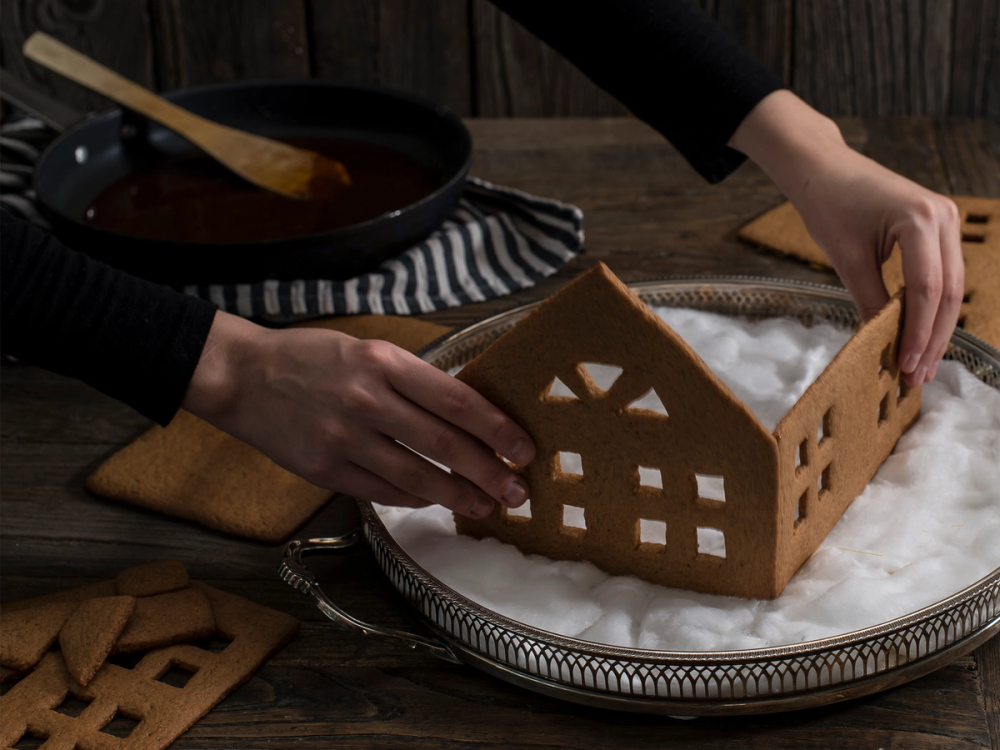 Person som setter sammen et pepperkakehus på et fat med mel eller sukker, med en stekepanne i bakgrunnen.