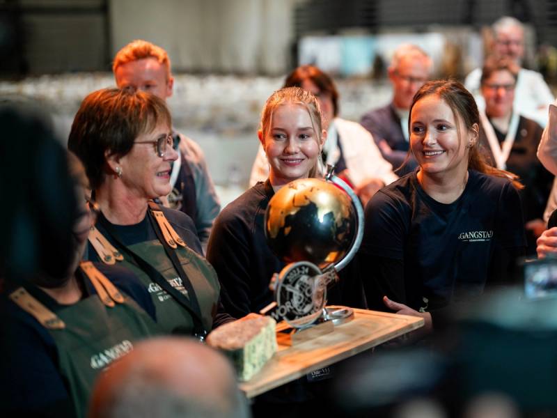 En gruppe mennesker samles rundt en pokal og en medalje under en utstilling eller premieutdeling, med flere personer i bakgrunnen.