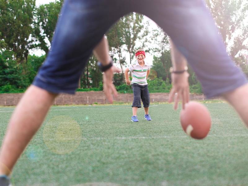 Barn som spiller amerikansk fotball på en gresslette med voksne i forgrunnen og trær i bakgrunnen.