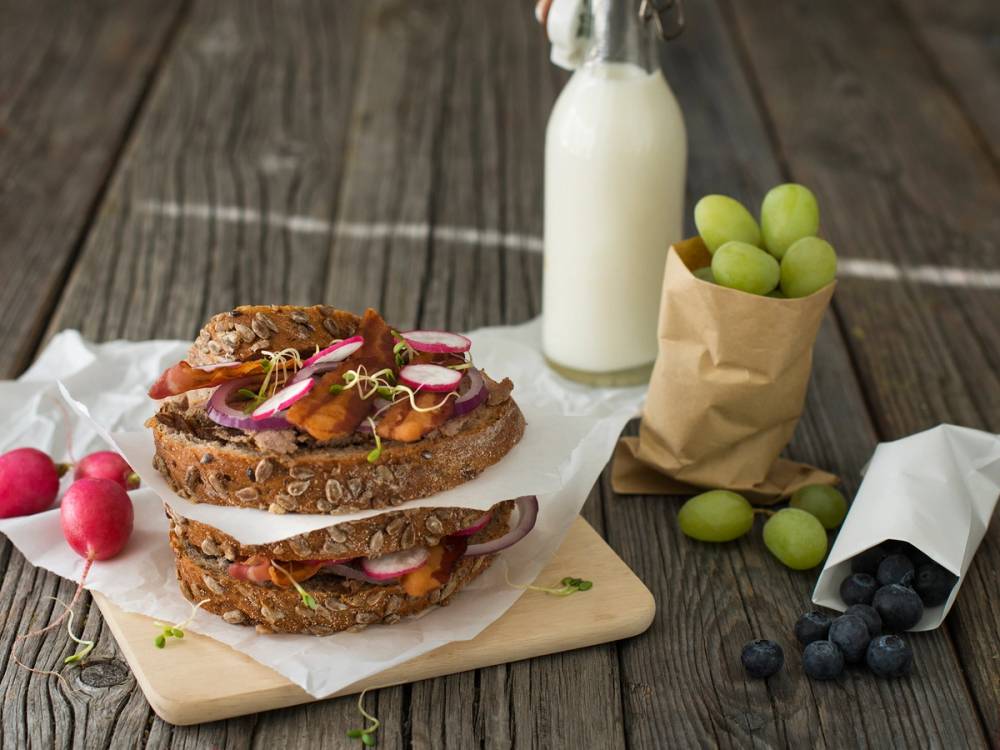 Grov leverpostei med bacon og tynne skiver av radiser og agurker på et brød, med en flaske melk og en papirpose med grønne druer i bakgrunnen.