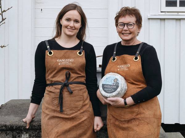 To personer står side om side foran en hvit bygning, begge iført brune forkle og holder en osteklokke, uten å vise ansiktene tydelig.