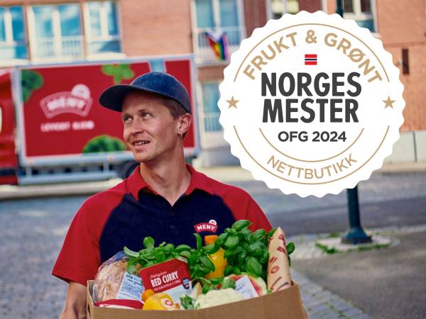 En person i rød og blå uniform holder en eske med frukt og grønt under Norges Mester 2024, med en logo for NM frukt og grønt på skjorten og en stor logo for arrangementet i bakgrunnen.