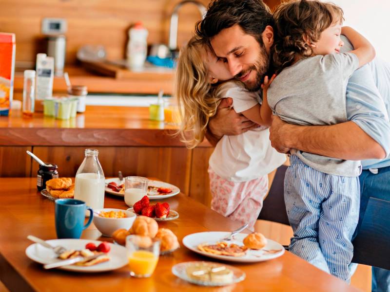 En voksen holder en liten gutt i armene mens de står ved kjøkkenbordet dekket med frokostmat, som egg, brød og melk.
