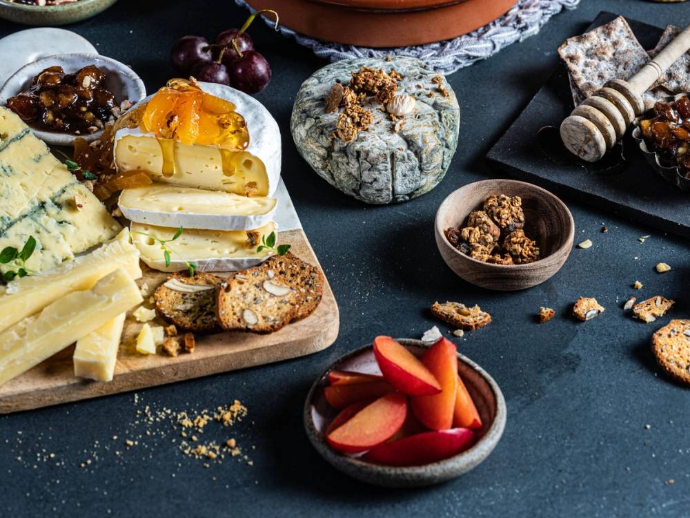 Et ostefat med ulike typer ost, kjeks, frukt og nøtter på et mørkt bord.