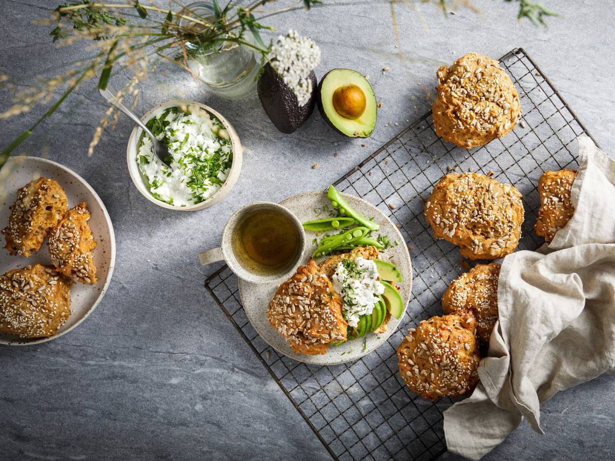 Rundstykker med havregryn på en avkjølt rist, omgitt av en skål med smør, en kopp med te, og et halvt avokado med et egg, på en grå overflate.