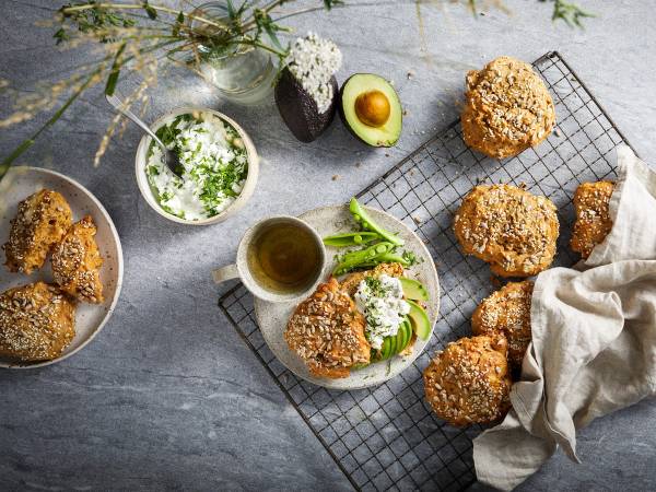 Rundstykker med havregryn på en avkjølt rist, omgitt av en skål med smør, en kopp med te, og et halvt avokado med et egg, på en grå overflate.