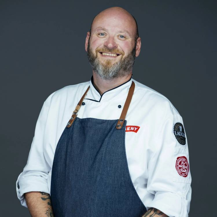 En person iført hvit kokkejakke og blå forkle, med armbånd og tatoveringer på armene, står mot en mørk bakgrunn.