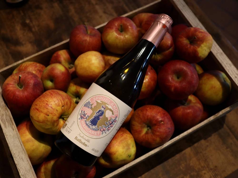 En flaske med eplesider plassert blant flere epler i en eske, på et treverk.