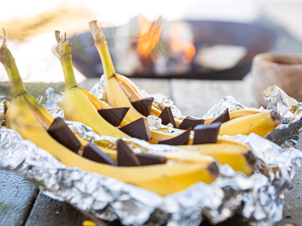 Bananer som er skåret opp og pakket inn i aluminiumsfolie, klare til grilling, med en grill i bakgrunnen.