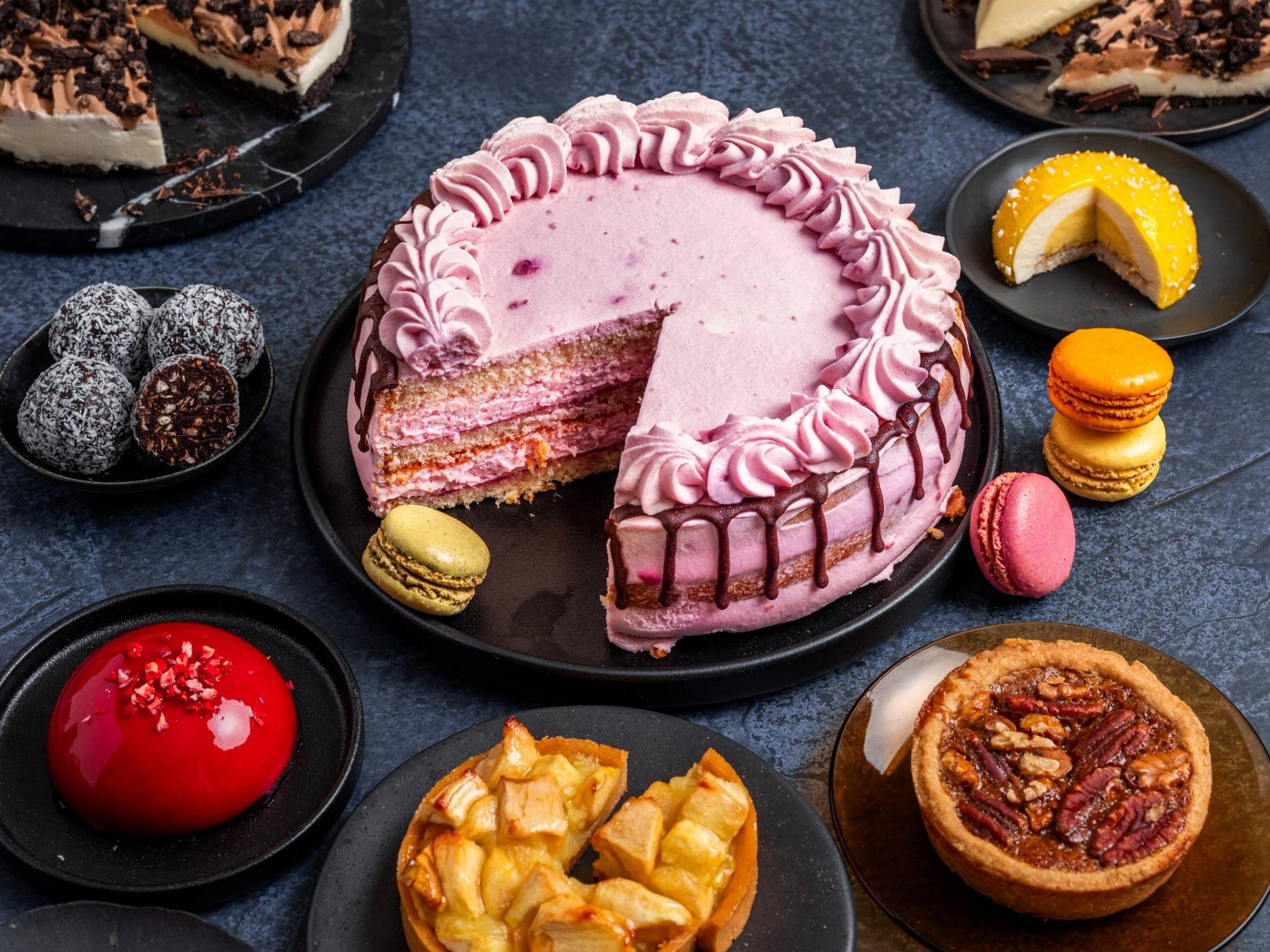 En kake med rosa krem og flere lag, omgitt av små kaker og desserter på en svart tallerken, på et mørkt bord.