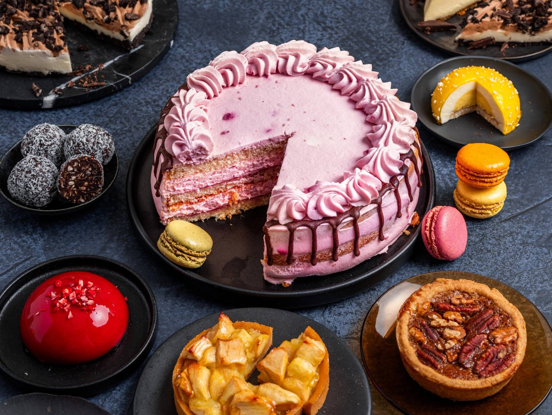 En kake med rosa krem og flere lag, omgitt av små kaker og desserter på en svart tallerken, på et mørkt bord.