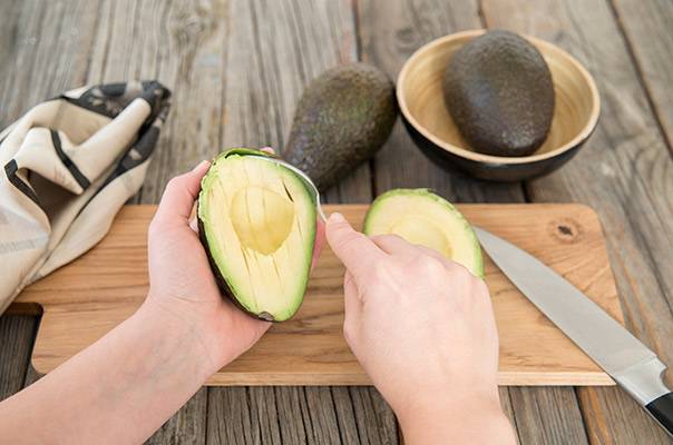 Person skreller en avocado på et skjærebrett med en kniv, mens to hele avokadoer ligger i en skål ved siden av.