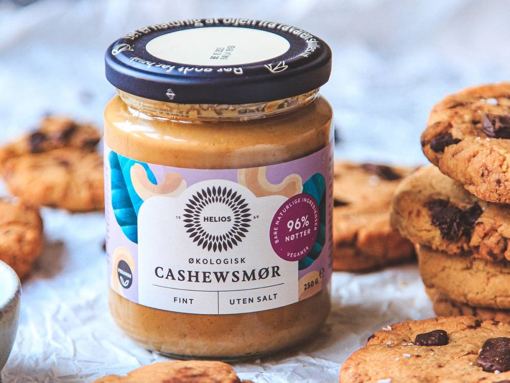 En glasskrukke med cashewsmør omgitt av sjokoladecookies med cashewsmør i bakgrunnen.