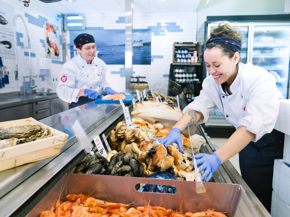 To ansatte i fiskebutikk håndterer fersk sjømat i en fiskedisk, med en rekke fisk og skalldyr foran seg.