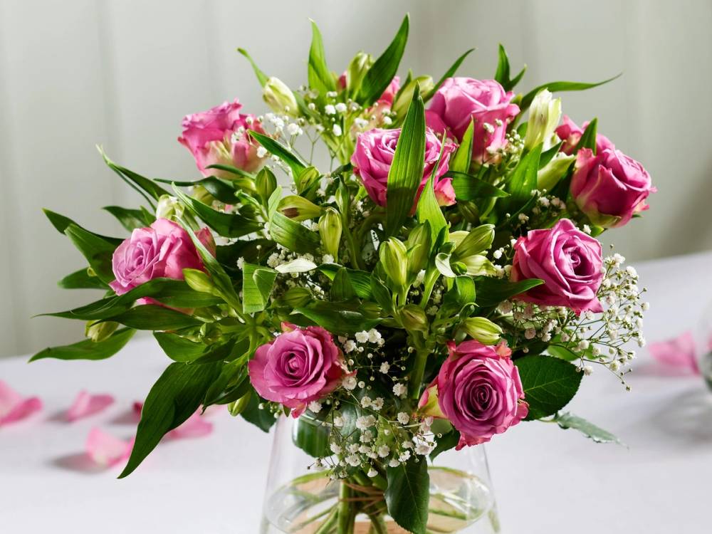 En bukett med rosa roser og hvit blomster i en glassvase, plassert på et bord.