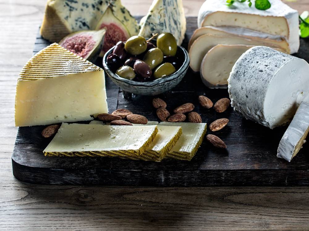 Utsnitt av en ostetallerken med ulike oster, grønne og svarte oliven, og noen nøtter på en treplate.